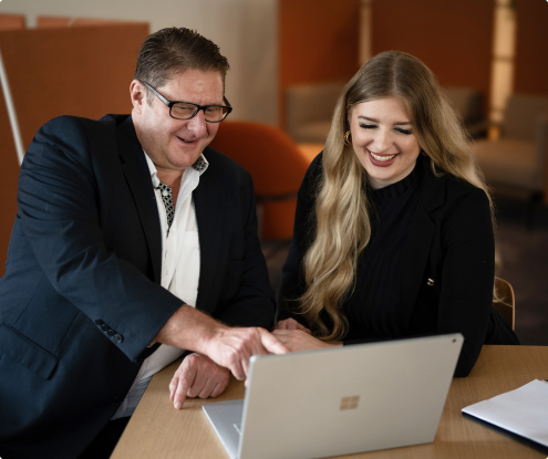 Zwei Personen in Geschäftskleidung schauen lächelnd auf einen Laptop und sind im Büro an einem Rekrutierungsprojekt beteiligt.