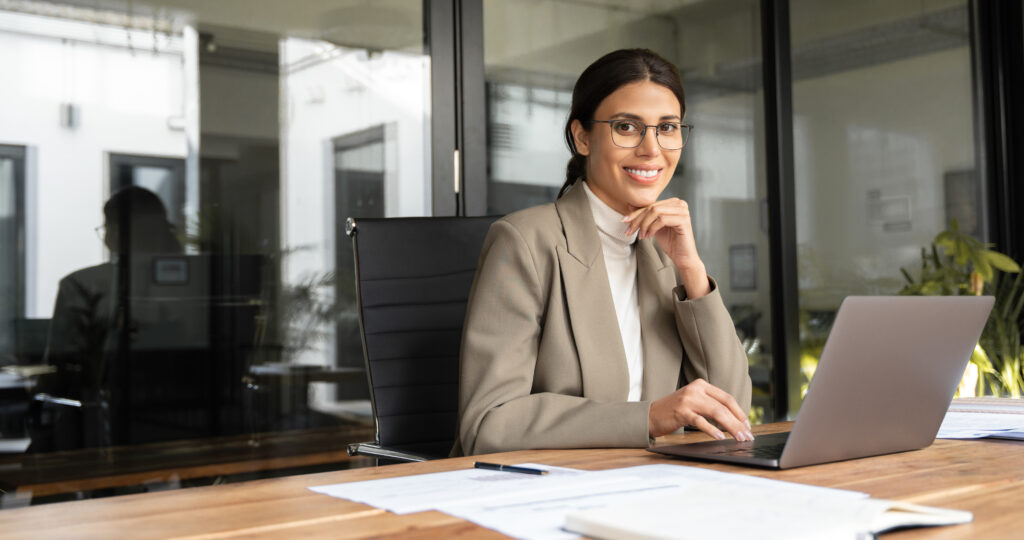 Businessfrau am Schreibtisch, arbeitet an Laptop und organisiert Bewerbungsunterlagen für eine erfolgreiche Bewerbung, großes Fenster im Hintergrund.
